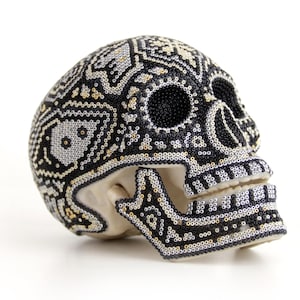 Puede incluir: Una calavera decorativa con intrincados abalorios en negro, plata y oro. La superficie de la calavera está cubierta con un patrón detallado, con cuentas negras que delinean las cuencas de los ojos y la boca. Un artículo único con temática del Día de Muertos.