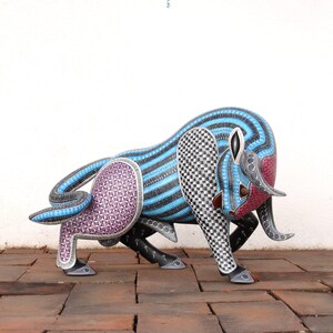 May include: A hand-carved wooden bull sculpture with intricate geometric patterns in shades of blue, black, white, and purple. The bull is standing on a brick surface.