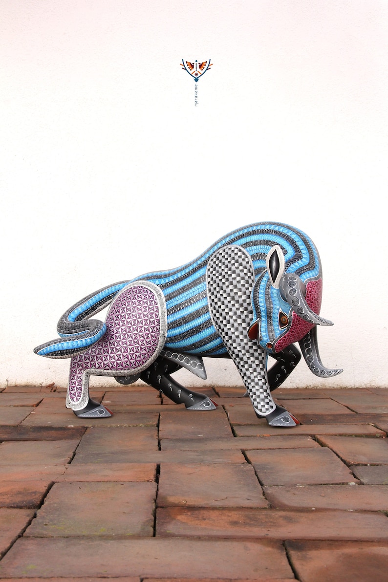 May include: A hand-carved wooden bull sculpture with intricate geometric patterns in shades of blue, black, white, and purple. The bull is standing on a brick surface.