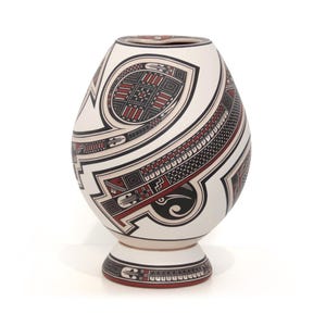 Puede incluir: Un jarrón de cerámica blanca con cuerpo redondeado y base de pedestal. El jarrón presenta intrincados diseños geométricos en negro, rojo y gris. Los patrones incluyen líneas, formas y motivos estilizados, creando una pieza visualmente llamativa.