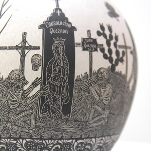 May include: Black and white ceramic sphere with a detailed carving of a graveyard scene. The carving includes skeletons, crosses, a tombstone with the text "Consolacion Quezada", and a figure of the Virgin Mary.