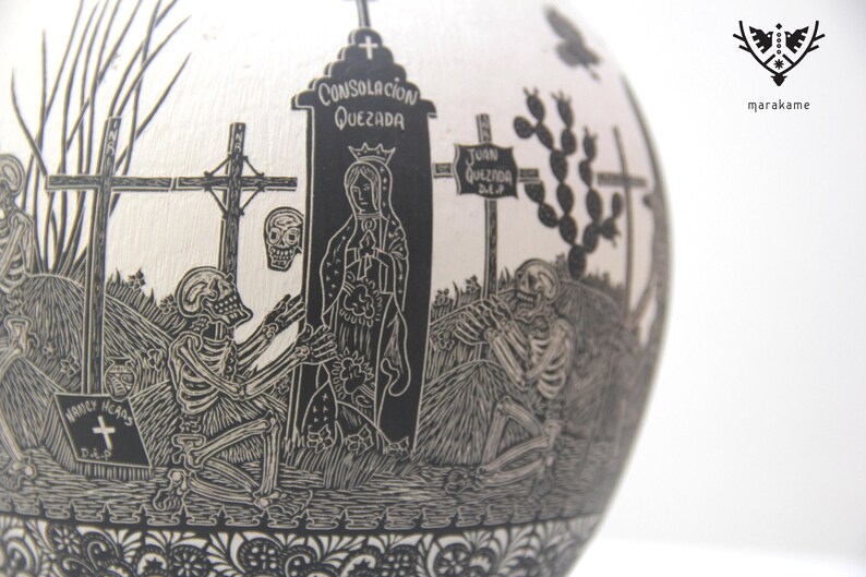 May include: Black and white ceramic sphere with a detailed carving of a graveyard scene. The carving includes skeletons, crosses, a tombstone with the text "Consolacion Quezada", and a figure of the Virgin Mary.
