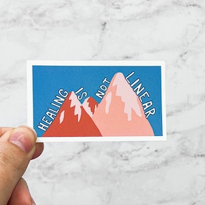 May include: A blue background with two pink and red mountains. The text "HEALING IS NOT LINEAR" is written on the mountains.