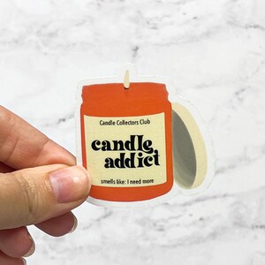 Puede incluir: Una pegatina transparente con un diseño de tarro de vela. El tarro es naranja y tiene el texto "Candle Collectors Club" y "candle, addict" en negro. El texto "smells like: I need more" también está en negro.