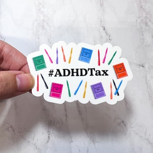 May include: A white sticker with the text "#ADHDTax" and illustrations of colorful pens and notebooks. The notebooks are green, blue, orange, pink, and purple. The pens are various colors, including pink, purple, yellow, red, and green.