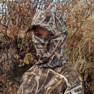 K&ouml;nnte beinhalten: Eine Person tr&auml;gt eine Tarnjacke und eine Tarnkappe mit einer Gesichtsmaske. Die Person steht vor einem Feld mit hohem Gras.