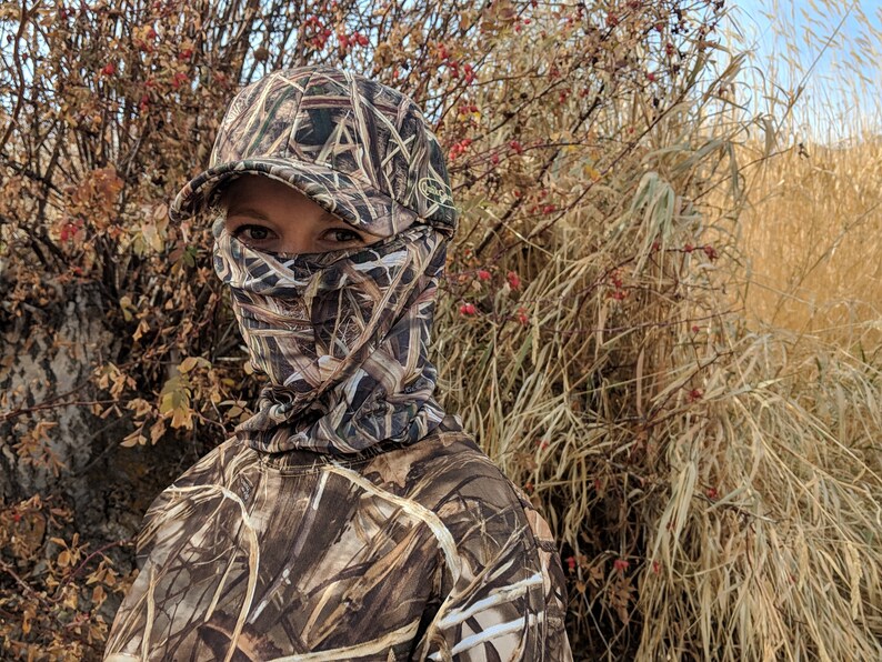 K&ouml;nnte beinhalten: Eine Person tr&auml;gt eine Tarnjacke und eine Tarnkappe mit einer Gesichtsmaske. Die Person steht vor einem Feld mit hohem Gras.