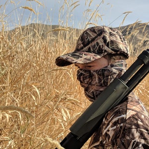 K&ouml;nnte beinhalten: Eine Person in Tarnkleidung und einer Tarnkappe mit dem Schriftzug "Duck Commander". Die Person h&auml;lt eine Schrotflinte.