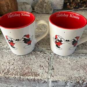 May include: Two white ceramic mugs with red rims. Each mug features a black and white illustration of Mickey Mouse and Minnie Mouse kissing. The text "Holiday Kisses" is printed in red on the front of each mug.