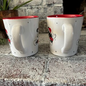 May include: Two white ceramic mugs with red rims. Each mug features a cartoon image of Mickey Mouse and a friend wearing Santa hats. The mugs have a white background with small black stars.