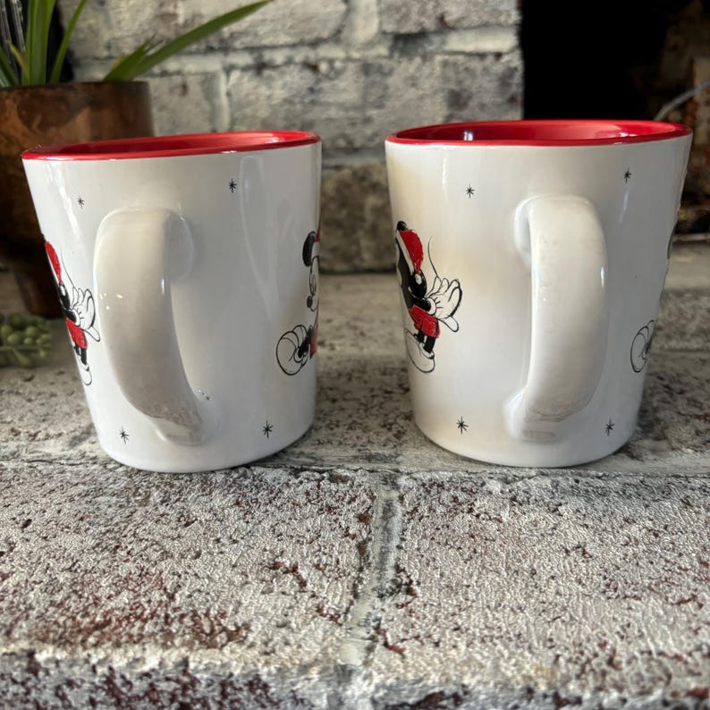May include: Two white ceramic mugs with red rims. Each mug features a cartoon image of Mickey Mouse and a friend wearing Santa hats. The mugs have a white background with small black stars.