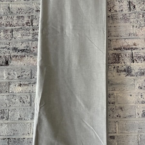 May include: A light gray rectangular tablecloth with a subtle texture. The tablecloth is hanging from a rod and is against a brick wall.