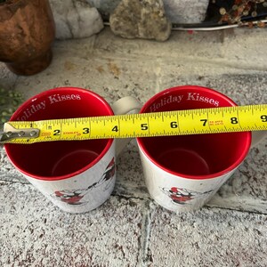 May include: Two red and white ceramic mugs with the text "Holiday Kisses" on the side. The mugs are being measured with a yellow tape measure.