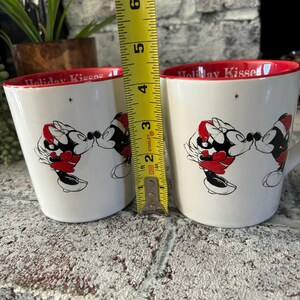May include: Two white ceramic mugs with red rims and a black and white illustration of Mickey Mouse and Minnie Mouse kissing. The mugs have the text "Holiday Kisses" on the front. A measuring tape is in the background.