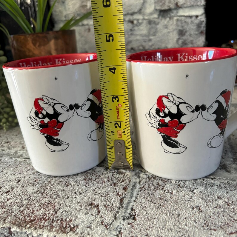May include: Two white ceramic mugs with red rims and a black and white illustration of Mickey Mouse and Minnie Mouse kissing. The mugs have the text "Holiday Kisses" on the front. A measuring tape is in the background.