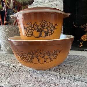 Może przedstawiać: Zestaw dwóch brązowych misek Pyrex do mieszania z wzorem owoców i winorośli. Mniejsza miska to przykryta naczynie do zapiekania.