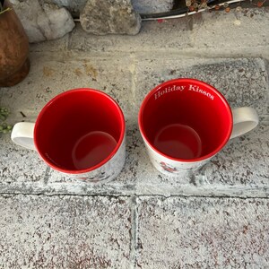 May include: Two white ceramic mugs with red interiors. One mug has the text "Holiday Kisses" printed on the side.