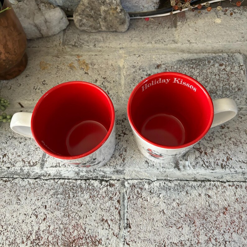 May include: Two white ceramic mugs with red interiors. One mug has the text "Holiday Kisses" printed on the side.