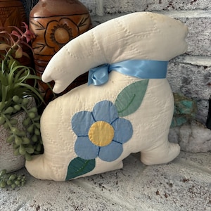 May include: A white fabric bunny with a blue flower and green leaves on its side. The bunny has a blue ribbon tied around its neck.