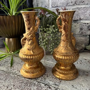 Puede incluir: Dos jarrones dorados antiguos con detalles florales y asas. Los jarrones tienen un acabado texturizado y brillante y una base bulbosa. El fondo incluye vegetación y una pared de ladrillo.