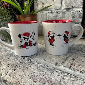 May include: Two white ceramic mugs with red rims. One mug features a black and white illustration of Mickey Mouse and Minnie Mouse dressed in Santa hats. The other mug features a black and white illustration of Mickey Mouse and Minnie Mouse kissing. The text "Holiday Kisses" is printed on the top of the mug.