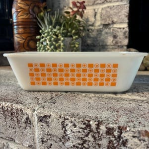 Peut inclure: Un plat de cuisson rectangulaire en verre blanc orné d'un motif orange répétitif composé de carrés pleins et de cercles avec des points centraux. Le plat présente une finition lisse et opaque ainsi qu'un large bord évasé, offrant un design épuré.