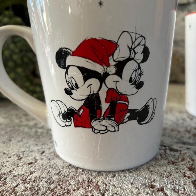 May include: White ceramic mug with a Christmas-themed design featuring Mickey Mouse and Minnie Mouse in Santa hats. The design is in black and white with red accents.
