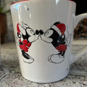 May include: White ceramic mug with a red rim featuring a black and white illustration of Mickey Mouse and Minnie Mouse kissing. Minnie is wearing a red Santa hat.
