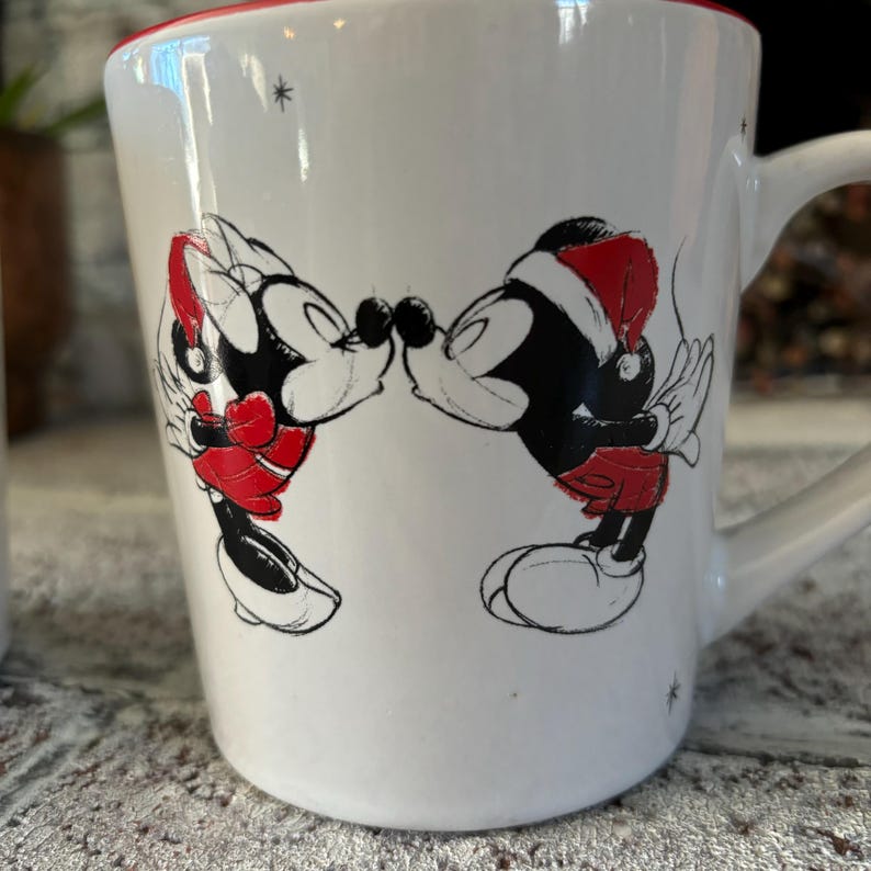 May include: White ceramic mug with a red rim featuring a black and white illustration of Mickey Mouse and Minnie Mouse kissing. Minnie is wearing a red Santa hat.
