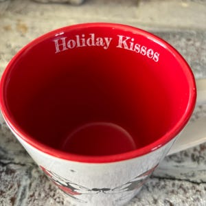 May include: White ceramic mug with a red rim and a black and white illustration of Mickey Mouse and Minnie Mouse kissing. The inside of the mug is red and has the text "Holiday Kisses" printed in white.