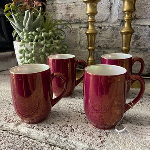 May include: Set of four burgundy ceramic mugs with iridescent glaze. The mugs have a rounded shape and a handle on the side. The mugs are sitting on a light gray surface.