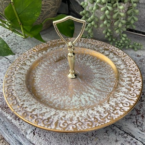 Puede incluir: Una bandeja de servir dorada con un diseño de remolino blanco y dorado y un asa dorada. La bandeja tiene un borde decorativo con un patrón de remolino.