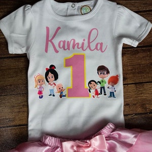 May include: White t-shirt with a pink number one and the name Kamila printed on it. The shirt features a cartoon image of a family of characters. The shirt is paired with a pink tulle skirt.