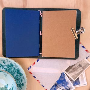 May include: An open, dark green leather journal with a blue and a tan page, held together with pink string. A silver clip holds the pages together. A floral teacup and saucer, envelopes, and postcards are nearby.