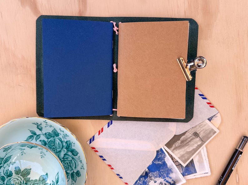May include: An open, dark green leather journal with a blue and a tan page, held together with pink string. A silver clip holds the pages together. A floral teacup and saucer, envelopes, and postcards are nearby.