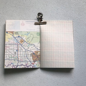 May include: Open notebook with a map of Santa Clarita, California, and graph paper pages. A silver binder clip holds the notebook open. The map side shows roads, parks, and the city name. The graph paper has red and blue lines.