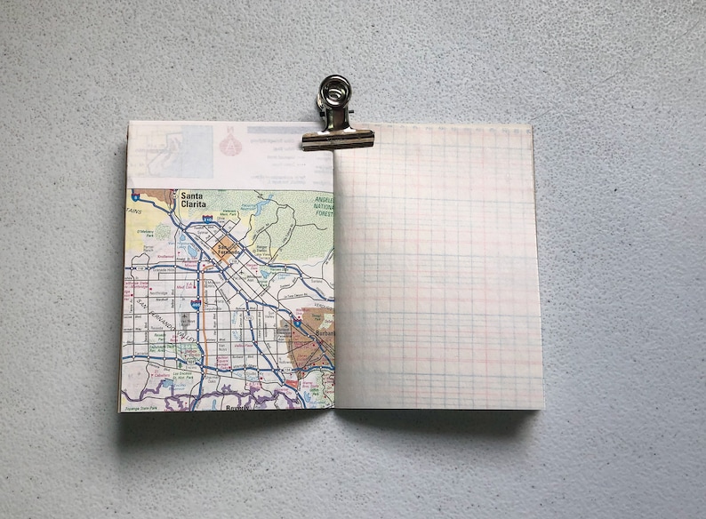 May include: Open notebook with a map of Santa Clarita, California, and graph paper pages. A silver binder clip holds the notebook open. The map side shows roads, parks, and the city name. The graph paper has red and blue lines.