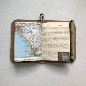 May include: A small, open notebook with a map of San Diego, California, and a dictionary page. The notebook is held by a metal clip on a brown leather-like backing. A black pen is resting on the right side.