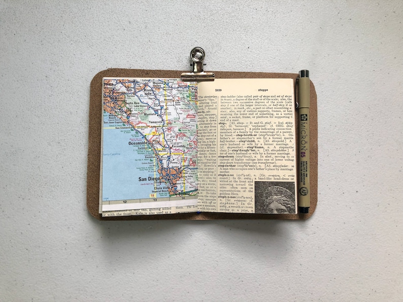 May include: A small, open notebook with a map of San Diego, California, and a dictionary page. The notebook is held by a metal clip on a brown leather-like backing. A black pen is resting on the right side.