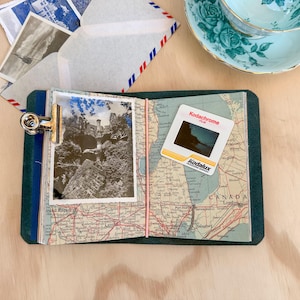 May include: A travel-themed journal with a vintage map design, holding a black and white photograph of a castle. A Kodachrome slide and a teacup are also present. The journal is secured with a metal clip and elastic band.