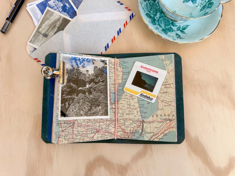 May include: A travel-themed journal with a vintage map design, holding a black and white photograph of a castle. A Kodachrome slide and a teacup are also present. The journal is secured with a metal clip and elastic band.