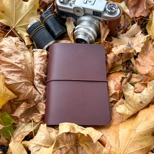Könnte beinhalten: Ein weinrotes, ledergebundenes Notizbuch mit elastischem Verschluss, eine Vintage-Kamera und zwei schwarze zylindrische Etuis liegen auf einem Bett aus Herbstblättern. Die Kamera ist silberfarben mit einem braunen Lederetui.