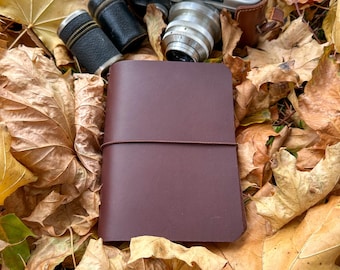 May include: A burgundy leather-bound notebook with an elastic closure, a vintage camera, and two black cylindrical cases rest on a bed of autumn leaves. The camera is silver with a brown leather case.