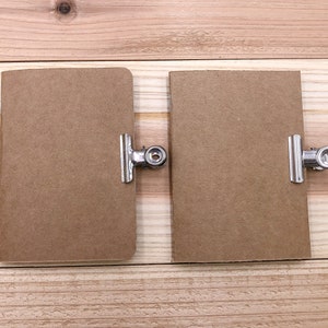 May include: Two small, rectangular brown notebooks with rounded corners are displayed side-by-side. Each notebook is held by a silver metal binder clip. The notebooks are on a light brown wooden surface.