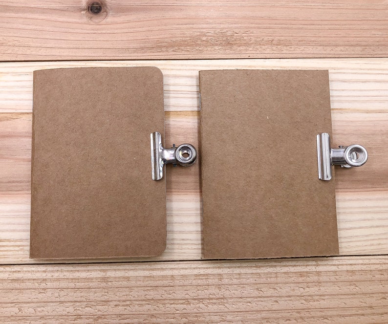 May include: Two small, rectangular brown notebooks with rounded corners are displayed side-by-side. Each notebook is held by a silver metal binder clip. The notebooks are on a light brown wooden surface.