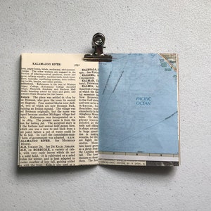 May include: A vintage map page with the Pacific Ocean visible, clipped to a dictionary page with the text "Kalamazoo River" using a silver binder clip. The pages are on a white surface.