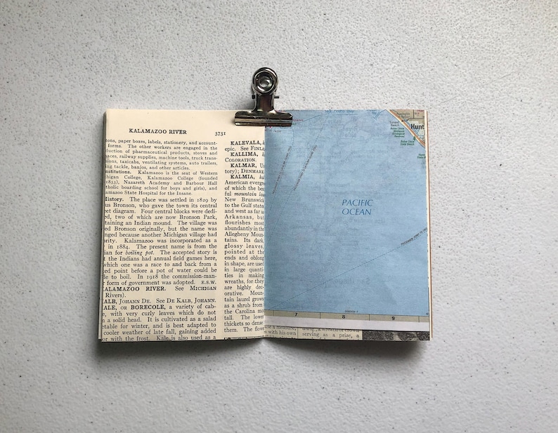 May include: A vintage map page with the Pacific Ocean visible, clipped to a dictionary page with the text "Kalamazoo River" using a silver binder clip. The pages are on a white surface.