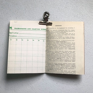 May include: An open book with a page featuring a green and white diagram and the other page with dictionary text. A silver binder clip holds the pages together. The book is on a light gray surface.