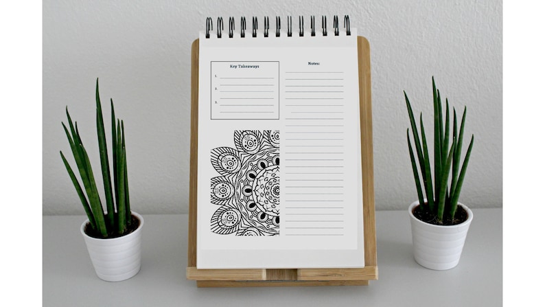 Puede incluir: Un cuaderno blanco de espiral con un dise&ntilde;o de mandala en blanco y negro en la p&aacute;gina izquierda. La p&aacute;gina derecha tiene una secci&oacute;n para "Puntos clave" y una secci&oacute;n para "Notas". El cuaderno est&aacute; en un soporte de madera y hay dos plantas en maceta a cada lado del cuaderno.