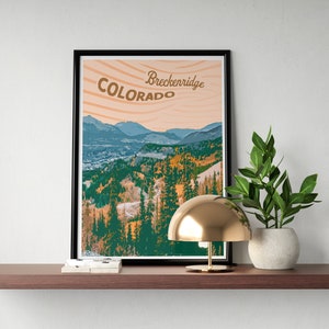 Op de afbeelding: Een ingelijste print met een berglandschap in Colorado, met de tekst "Breckenridge Colorado". De print staat op een houten plank, samen met een gouden lamp en een plant in een pot.